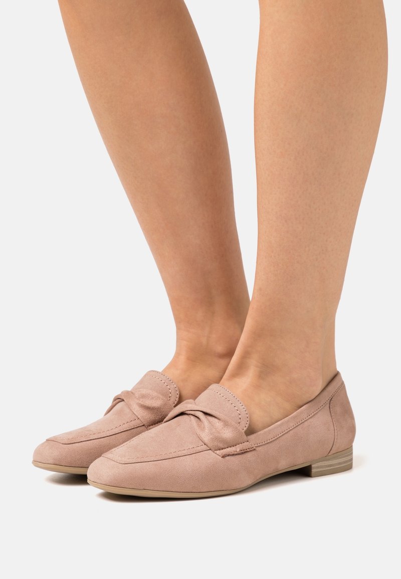 Paire de mocassins en daim rose clair avec un détail de nœud, portés pieds nus sur un fond clair uni.