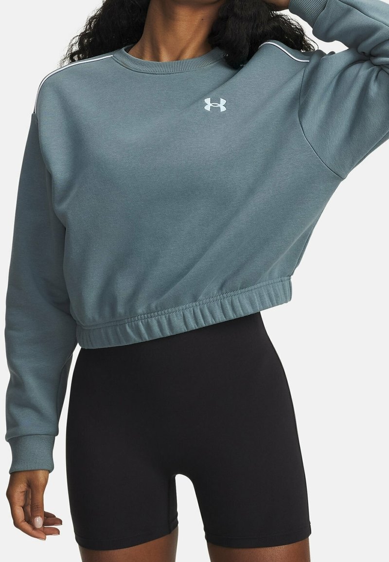 Femme portant un sweat-shirt Under Armour court bleu et un cuissard noir ajusté, posant avec un bras levé derrière la tête.