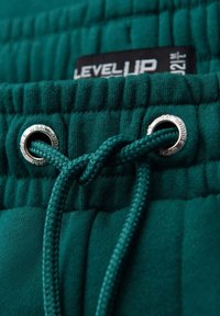 Cintura de sudadera de algodón verde con cordón y ojales plateados. El tejido tiene un acabado texturizado y una etiqueta que dice "LEVEL UP."