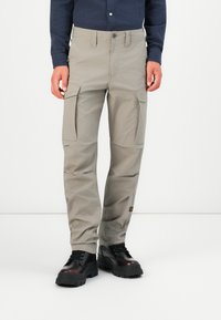 Beige cargobroeken met meerdere zijzakken, gemaakt van lichtgewicht stof, gecombineerd met zwarte enkellaarsjes en een donkerg blauwe knop-down shirt.