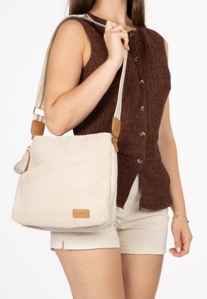 Frau trägt ein ärmelloses, braunes, knöpferbesetztes Oberteil und beige Shorts, hält eine große, strukturierte, beige Schultertasche mit braunen Akzenten.