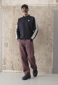 Crna dukserica s bijelim prugama, smešaste hlače i crne patike. Sadrži Adidas logo na grudima. Stoji naslonjen o teksturirani zid.