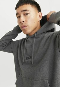 Sudadera gris hecha de un tejido suave, con un bolsillo tipo canguro, cordones y el logo "JACK & JONES" en negro en el pecho. Textura lisa.