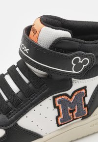 Sneaker high-top in pelle nera con accenti bianchi, caratterizzato da una patch "M" grigia e arancione, una fascetta in velcro e dettagli in mesh traspirante.