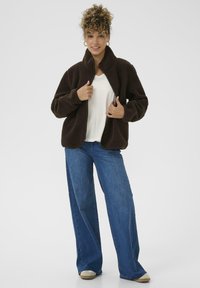 Giacca in pile marrone con colletto alto, chiusura con zip e due tasche laterali, abbinata a jeans a gamba larga blu e scarpe chiare.
