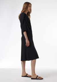 Vestido negro de jersey con escote en V drapeado y mangas cortas estilo murciélago, hasta la rodilla, combinado con sandalias minimalistas negras. Textura suave en toda la prenda.