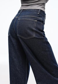 Tmavě modré džínové kalhoty s volnějším střihem, vybavené dvojitými zadními kapsami a kontrastním prošíváním. Hladká textura a široký střih nohavic.