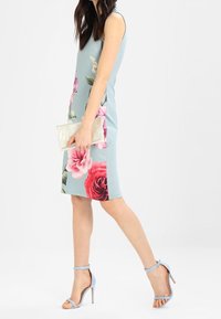 Femme marchant dans une robe sans manches bleu pâle à gros imprimé floral rose, tenant une pochette métallique et portant des sandales à talons hauts assorties bleu.