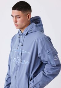 Giovane uomo che indossa una giacca con cappuccio blu con il logo "PXP" sul petto e sulla manica, che guarda di lato contro uno sfondo semplice.