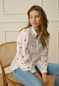 Jonge vrouw met lang haar die een wit blouse draagt met rode rozen- en hartmotieven, zittend op een houten stoel met een geweven zitting.