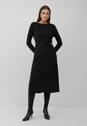 Woman standing, wearing a black long-sleeve midi dress with a side buckle, black tights, and black heeled ankle boots, against a plain background.
