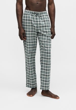 Mand står barfodet iført grå og hvide ternede pyjamabukser med snøre i taljen, forkortet til midt på torsoen.