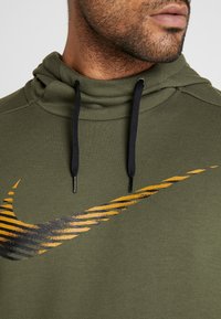 Sweat à capuche vert olive avec un col montant, doté de cordons de serrage noirs et d'un logo Nike ondulé diagonal orange et noir sur la poitrine.