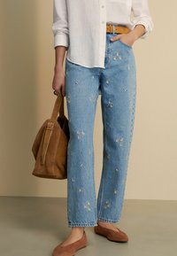 Jeans de mezclilla azul con bordados florales, corte relajado, dobladillo deshilachado, combinados con una camisa blanca de botones y un bolso de ante marrón.