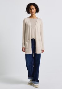 Gilet en tricot beige à ouverture frontale, avec des poignets côtelés et deux poches avant, associé à un haut léger et un jean bleu ample.