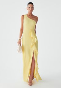 Yellow one-shoulder dress with a textured fabric, ruffle detail down the front, and a thigh-high slit. Paired with a white woven handbag.