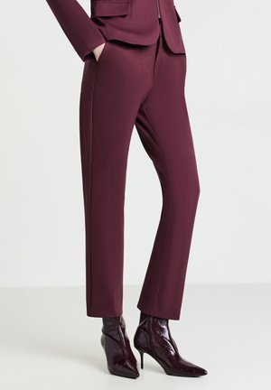 Femme portant un pantalon bordeaux ajusté et des bottines assorties à bout pointu et talons aiguilles, se tenant les mains dans les poches.