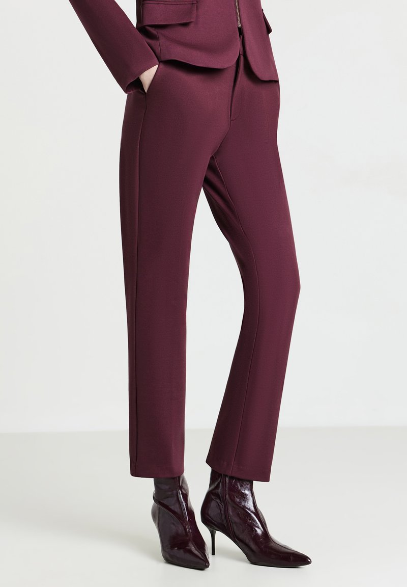 Donna che indossa pantaloni attillati color bordeaux e stivaletti coordinati con punta a punta e tacco a stiletto, in piedi con le mani nelle tasche.