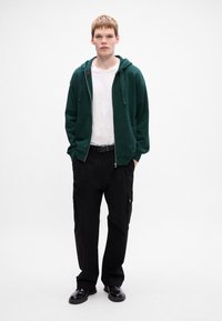 Grøn zip-up hoodie med en kængurulomme over en hvid t-shirt, parret med sorte cargo-bukser med sidelommer og sorte sko.