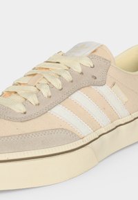 Κοντινή λήψη μπεζ αθλητικού παπουτσιού Adidas Samba με κρεμ κορδόνια, λευκές τρεις ρίγες, επενδύσεις από σουέτ και παχύ πάτο εκρού.