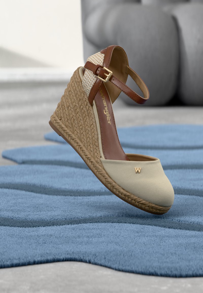 Μπεζ πλατφόρμα πέδιλο με πλεκτή φτέρνα από γιούτα, καφέ λουράκι αστραγάλου με αγκράφα και χρυσό έμβλημα "W" στη μύτη, τοποθετημένο σε μπλε χαλί.
