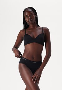 Reggiseno nero e slip abbinati, entrambi con una texture liscia. Il reggiseno ha spalline sottili e un logo discreto sulla vita degli slip.