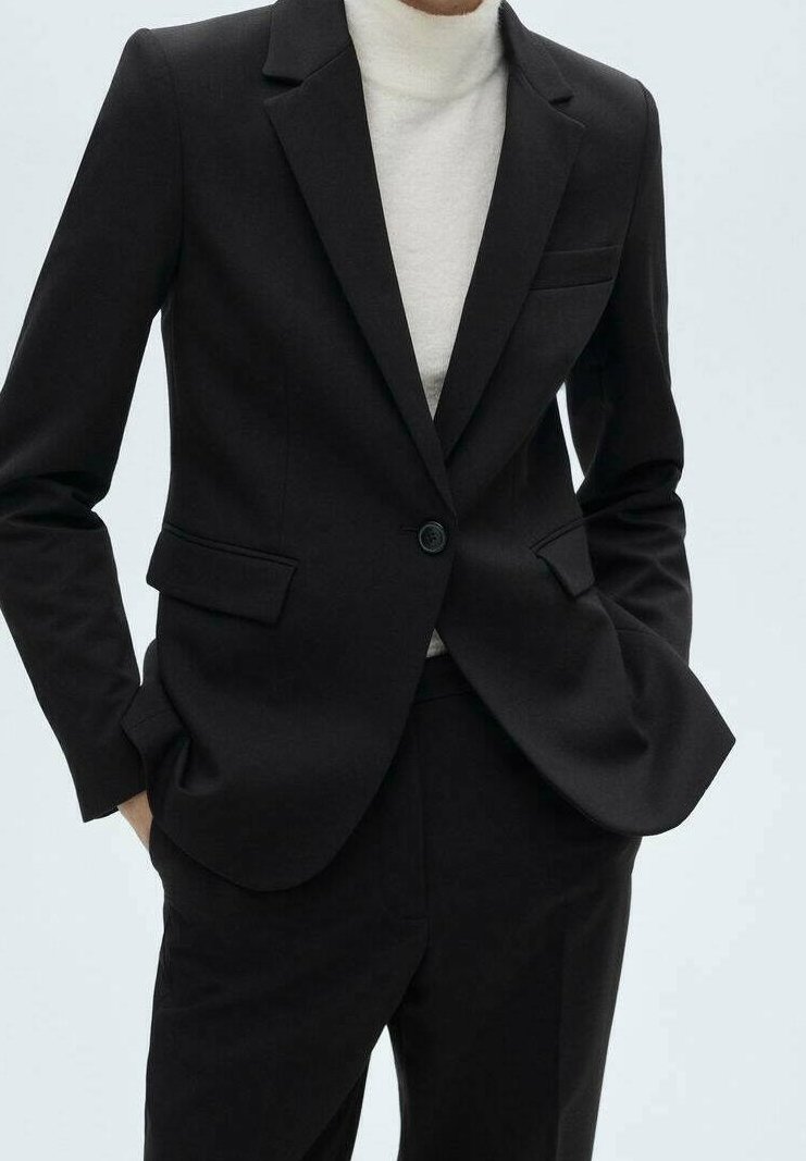 Personne portant un blazer noir à un bouton et un pantalon assorti, les mains dans les poches, associé à un haut à col roulé blanc.
