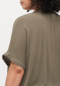 Olive green short-sleeve top in lightweight, textured fabric with a relaxed fit and rounded neckline, showcasing soft drape and subtle sheen.