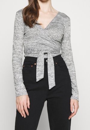 Top enveloppant gris en tissu tricoté texturé avec des manches longues, doté d'un décolleté en V profond et d'un détail de taille nouée. Associé à un pantalon noir taille haute.