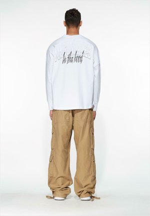 White long-sleeve shirt with large gray text on the back, paired with beige cargo pants featuring multiple pockets and a relaxed fit.
