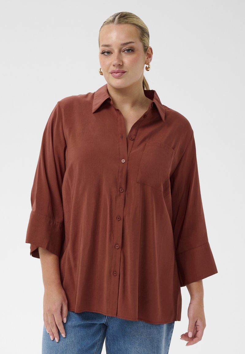 Femme aux cheveux blonds plaqués en arrière, portant des boucles d'oreilles en or, une chemise marron ample boutonnée et un jean bleu, debout devant un fond uni.