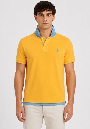 Homme portant un polo jaune avec un col bleu et un pantalon de couleur claire, debout sur un fond uni clair.