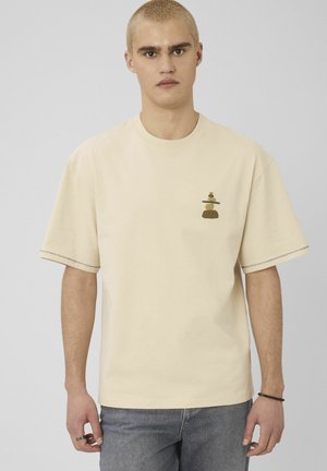 Jeune homme aux cheveux blonds courts portant un t-shirt beige ample avec un petit motif de pierres empilées équilibrées sur le côté gauche de la poitrine.