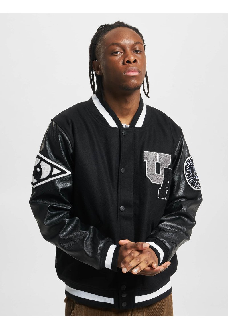 Veste varsity noire avec un corps en laine et des manches en cuir. Présente des rayures blanches sur le col et les poignets, ainsi que des écussons sur les bras et la poitrine.