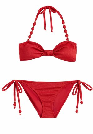 Femeie în bikini roșu, mergând desculță pe apă puțin adâncă la plajă, ținând o geantă țesută, cu palmieri și ocean în fundal.