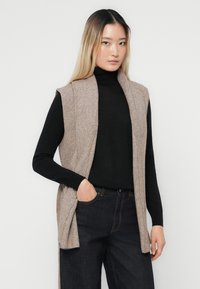 Beige gebreide vest met een geribbelde structuur, open voorkant en zonder mouwen, gedragen over een zwarte coltrui. Donkerdenim jeans met een klassieke pasvorm.