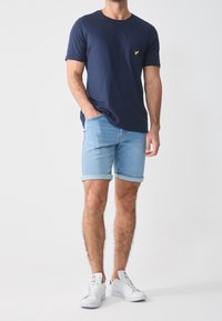 Námořnická modrá tričko s krátkým rukávem a kapsou na hrudi, ve spojení se světle modrými vyhrnutými džínovými šortkami a bílými teniskami. Jednoduchý design s minimálními akcenty.