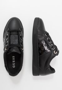 Černé tenisky Guess s lesklými vzorovanými bočními panely, černými tkaničkami a silnými texturovanými podešvemi, zobrazené z horního a bočního pohledu.