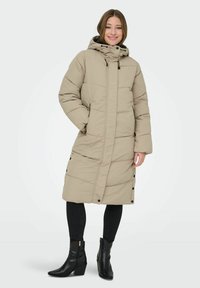 Abrigo puffer largo beige con capucha, con textura acolchada y botones en los laterales. Combinado con jeans ajustados negros y botas de tacón.