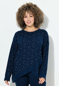 Frau mit lockigem Haar, die einen marineblauen Pullover mit Sternenmustern und marineblauen Hosen trägt, vor einem schlichten weißen Hintergrund steht.