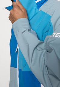 Osoba v modrošedém dešťovém kabátu s nápisem "RAIN DRY" na rukávu, která si upravuje límec rukou.