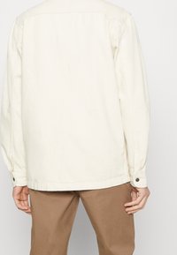 Beżowa kurtka z płótna o luźnym kroju, z mankietami na guziki i prostym dołem. Tył ma prosty wzór bez zdobień.