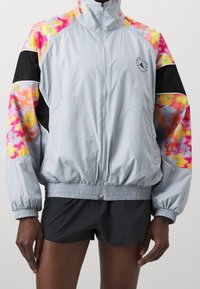 Chaqueta de cremallera gris claro con mangas de patrón tie-dye colorido, puños elásticos y cinturilla fruncida. Presenta un logo en el pecho.