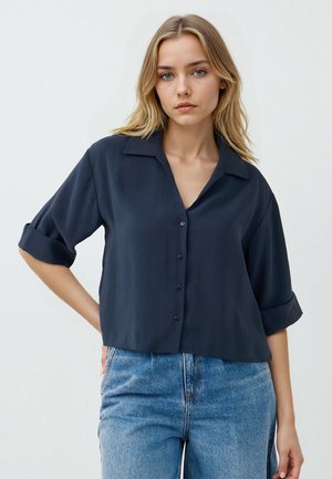 Marineblaue Bluse mit kurzen Ärmeln, Kragen, Knopfleiste und cropped Design. Glatte Textur, hochgerollte Ärmel und lockere Passform.