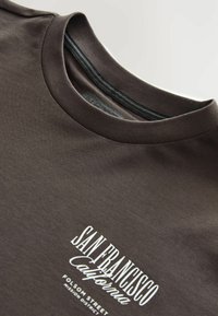Brun bomulds t-shirt med rund halsudskæring. Har hvidt trykt tekst "SAN FRANCISCO California FOLSOM STREET" i varierende skrifttyper.