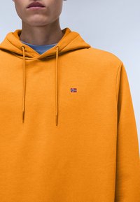 Orange hoodie made of textured material with a drawstring hood and a small embroidered flag on the chest.