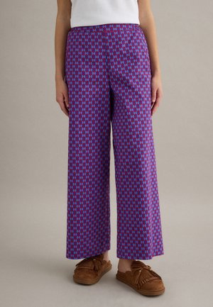 Persoon draagt een broek met wijde pijpen in een blauw-rood geometrisch patroon, een wit shirt en bruine instapschoenen met kwastjes, staand op een grijze achtergrond.