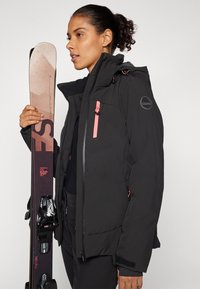 Giacca da sci nera con dettagli rosa, tasche con zip e cappuccio regolabile. Modello che tiene gli sci con attacchi neri e motivo rosa.