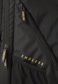 Juodas audinio maišas su keliais užtrauktukais, kiekvienas turintis juodus vilkimo užtrauktukus su geltonu tekstu "ENDLESS", ir šoninė tinklinė kišenė.
