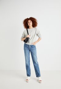 Sweatshirt gris, jeans bleu droit, et talons ouverts blancs. Sac à main noir avec une bandoulière et cheveux bouclés rouges. Fond uni.